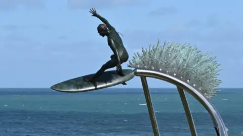 Jonathan Start A statue of a surfer on a surf board suspended on a steel pole with the sea behind it. Behind the surfer is a bronze wave symbol