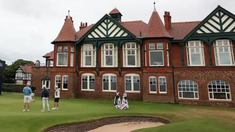 Getty Images Golfers practise on the 18th green prior to the AIG Women's Open 2026 at Royal Lytham & St. Annes. The club house is a two-storey building built from red bricks and with mock-Tudor features.
