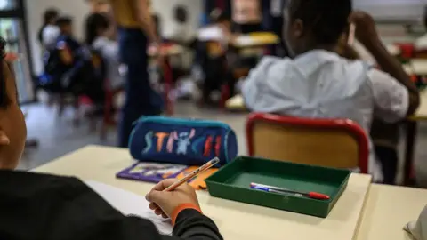 Getty Images A close-up view from a desk shows a child holding a pen in their right hand. A blue pencil case and a small green tray containing pens sit on the desk. Other children are seated ahead but appear out of focus in a classroom learning environment.