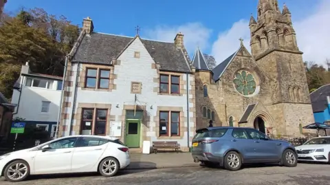 The hall is a small, two-storey building with a green door and square windows. It is next to an old church. Three cars, one blue and two white, are parked on the street outside the buildings.