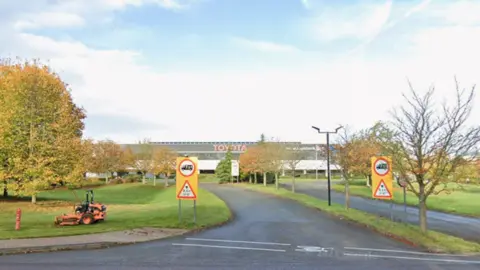 Two yellow signs in front of a patch of grass, which is in front of the Toyota manufacturing plant