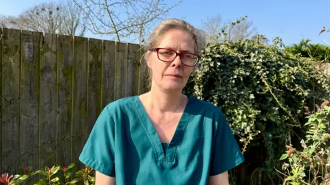 Becki Bowden / BBC Michaela is wearing a glasses and a green top. Her hair is tied back in a clip. She is stood in front of her garden fence and is looking at the camera. 