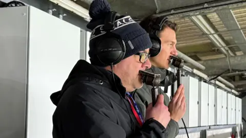 A man with glasses and a woolly hat holding a microphone to his mouth while standing on a gantry. Another man standing just behind him is also speaking into a microphone.