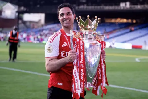 Getty Images Mikel Arteta smiles as he carries the Premier League trophy across the pitch, with stewards and fans visible in the background.