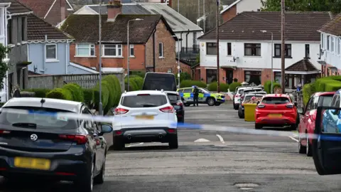 A row of multiple cars sit either side of a residential road. There are red-brick houses the run the length of the road. At the bottom of the road a blue and yellow police car sits behind blue and white police tape. A police officer in black uniform stands beside the car. 