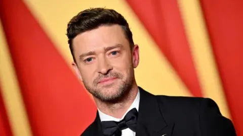 Justin Timberlake wearing a suit and bow tie in front of a yellow and red background.