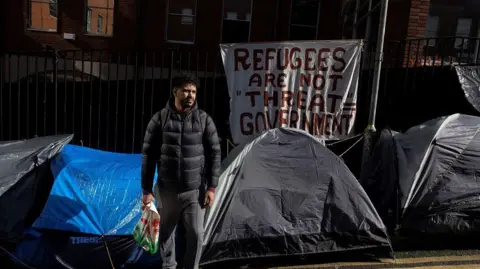 REUTERS/Clodagh Kilcoyne Description An Algerian homeless asylum seeker named Walim walks beside tents outside the International Protection Office (IPO) where hundreds of asylum seekers looking for accommodation have been sleeping on the streets for several months with more arriving every day, in Dublin, Ireland, March 14, 2024. REUTERS/Clodagh Kilcoyne TPX IMAGES OF THE DAY