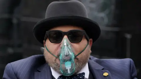 PA Media Nicholas Rossi wearing a pinstripe suit, black sunglasses, black hat and an oxygen mask. He has a grey beard.