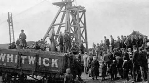 Whitwick Colliery Disaster Facebook Group A black and white photo, showing a coal wagon with the word Whitwick, on the side in large letters. It stands in front of a pit head structure.  A crown of men stand around it