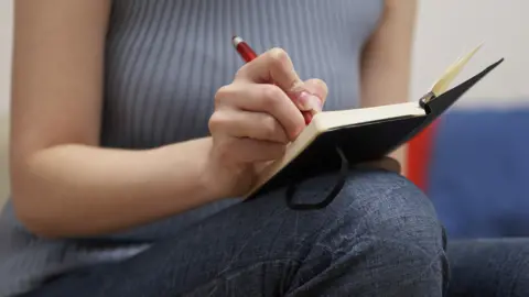 A close up of a woman's hand holding a pen as she writes in a black hard back notebook while sitting on a sofa.