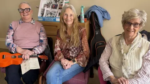 Three elderly women sat on chairs smile at the camera. One has a ukulele and music sheet on her lap.