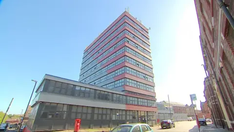 The Horizon building which is half-lit by sunshine. There are cars parked in the street and a red post box.