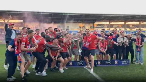 A football team in red shirts lift a trophy and spray champagne
