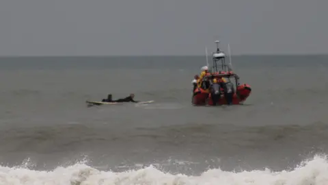 Neil Phillips Oscar is manoeuvring the man on his surfboard towards the RNLI inshore lifeboat.