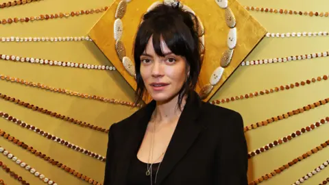 Getty Images Lily Allen pictured half-smiling wearing a black outfit against a stylised yellow background