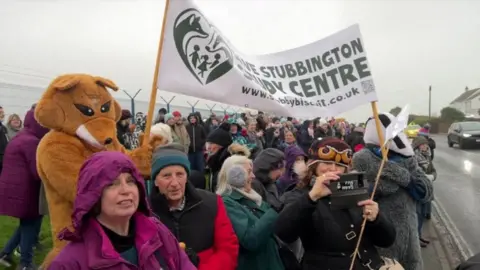 Hundreds of protesters standing on a footpath next to a busy road. Two people dressed as a fox and a badger are carrying a banner that says Save Stubbington Study Centre