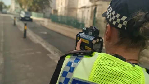 Devon and Cornwall Police The image shows a police officer standing at the side of a city street using a handheld speed detection device aimed at approaching traffic. The officer is wearing a high-visibility jacket with a blue-and-white chequered pattern and the word "POLICE" visible on the back, while a black-and-white chequered police cap is also visible.