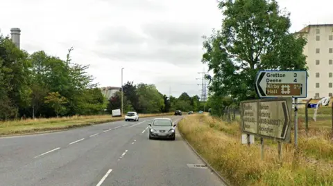 Google The A6116 in Corby showing cars and signage for the Gretton junction. There are industrial buildings and a power station chimney close to the road. It is a dull day.