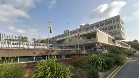 Google Three flagpoles outside a concrete council building. They fly the yellow and blue Ukraine flag, the Union flag and the red and white county flag