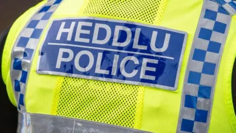 The back of an officer's yellow and blue vest. The word 'Police' and 'Heddlu' are in silver and in a blue box.