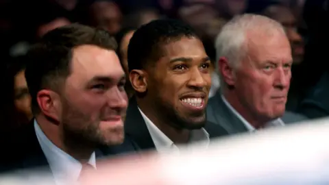 PA Media Eddie Hearn, Anthony Joshua and Barry Hearn smartly dressed and sitting ringside at a boxing fight.