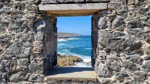 A beach and sea and cliffs and blue sky seen thought the window slot of an ruined stone building.