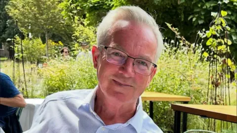 Ash Family An older person with light hair and glasses sits outdoors at a cafe-style garden table, surrounded by greenery and other people in the background.