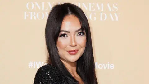 Sharon Gaffka has long dark hair and is wearing a black sequined dress. She is standing in front of an ivory wall.