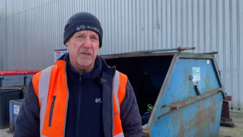 Manager Gary Booth wearing a hat and high vis stood in front of a blue skip 