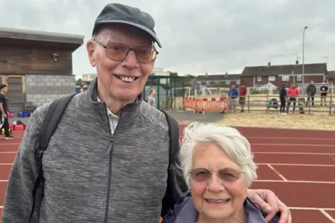 BBC / Scott Dalton The photo shows a man and a woman smiling at the camera. The man on the left has a grey jacket on. He's also wearing glasses and a grey cap. He has his arms on the shoulders of the woman, on the right. She is wearing a purple jacket and they are standing on a brown athletics track. There is also a grey brick building behind them. 