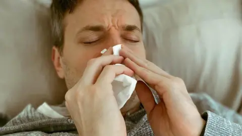 Getty Images Man with stubble wearing grey top, blowing nose with a white tissue.