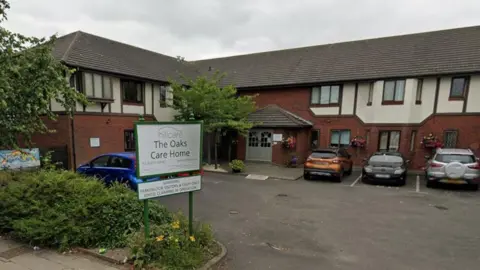 The Oaks Care Home. A green and white sign stands out of the front of the building outside the care park. The building is made of red brick with white sections around its windows. Several cars are parked in bays near the entrance.