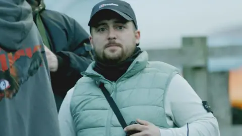 Connor Graham standing behind a metal barrier at a protest. He has a beard and is wearing a black baseball cap, light grey top and a bodywarmer.