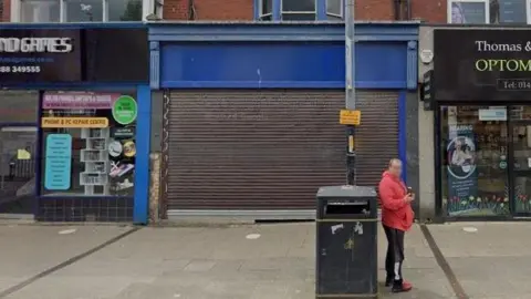 A Google Street View screenshot of the Screams Court 2.0 venue in Hartlepool. The unit has its shutter down and has a blue panel without a sign. It is flanked by shops. A man in a red hoodie and tracksuit bottoms is drinking a can outside.
