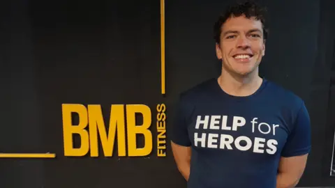 Rob Guest. He has short, dark, curly hair and is wearing a navy blue Help for Heroes t-shirt. He is standing against a black wall with a yellow BMB Fitness logo on it. He is smiling, looking directly at the camera. 