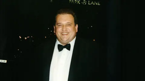 Family photo Phil is wearing a formal black suit with a white shirt and black bow tie. He is smiling at the camera. The background is completely dark. 