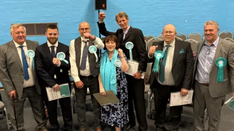 Seven Reform UK politicians are seen smiling and celebrating. They are wearing light blue rosettes and are stood against a blue wall.