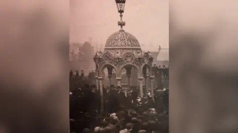 March and District Museum Black and white picture from 1912 of big crowds gathered around a domed monument complete with water drinking feature in the centre. The structure is ornate, with eight open sides with arches and eight pillars supporting the dome. It stands about 8m (25ft) high and has a Victorian looking gas lamp on the top.