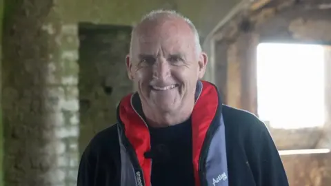 A man with short white hair smiling at the camera. He is wearing a black jumper and a red and grey life jacket. He is stood in an old brick fort.