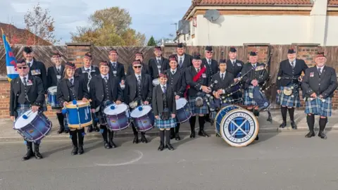 Newton Aycliffe Pipe Band The Newton Aycliffe Pipe Band before their performance at Remembrance Sunday. They are all in their formal dress with blue kilts and suits. They are carrying their drums and pipes.
