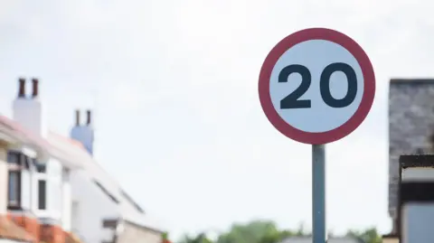 Red 20mph road sign with homes in the background.
