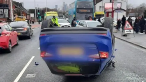 A blue Skoda Fabia resting on its head after crashing. The car is in the middle of a congested road. There is a bus in the foreground.