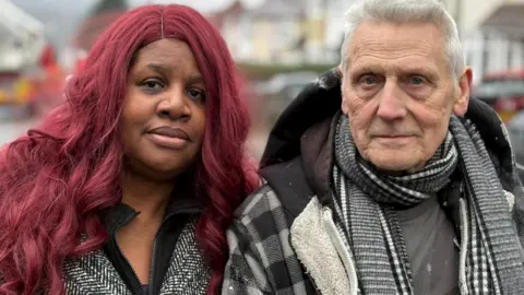 A woman and man stand side by side. Both wear winter coats and the man also wears a scarf. They are looking at the camera. 