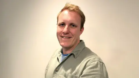 Steffan Evans A young man with short blonde hair, smiling and wearing a light brown shirt.