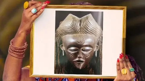 Samba Yonga from Women's History Museum of Zambia holds up a frame over her face showing a photograph of a sacred mask with Sona symbols etched on to its surface, each telling stories of women's significance, wisdom, and the vital knowledge they carried.