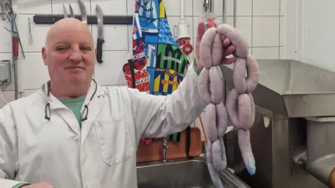 Dan Morgan A butcher holding up blue skinned-sausages inside a butchers back room 