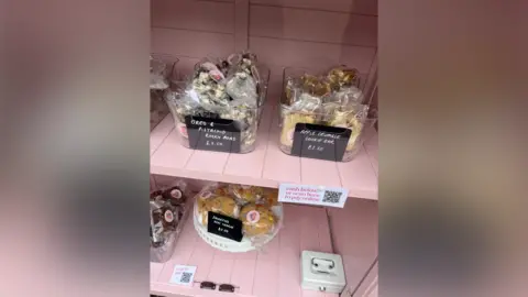 Susie White Pink wooden shelves have multiple plastic clear boxes on them containing packaged baked goods. 