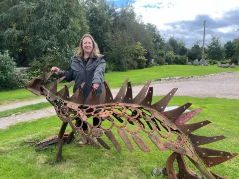Mujer respalda una gran escultura de metal oxidada de algún tipo de reptil o dinosaurio. Parece tener picos contundentes que corren a lo largo de su respaldo. En su mayor es más grande, es tan alto como el pecho de la mujer. 