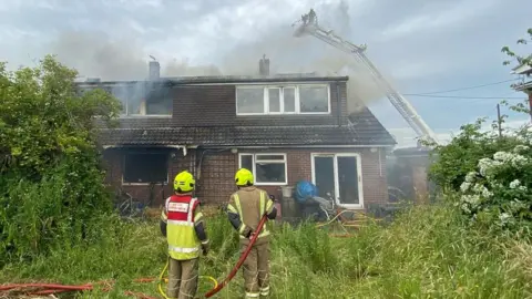 Essex Fire and Rescue Service Two firefighters standing in front of a house engulfed with smoke, with an aerial platform towering over it.