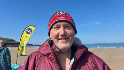 Paddy McMonagle with a grey beard standing on a beach in a purple dry robe who is wearing a  red and black rugby beanie hat